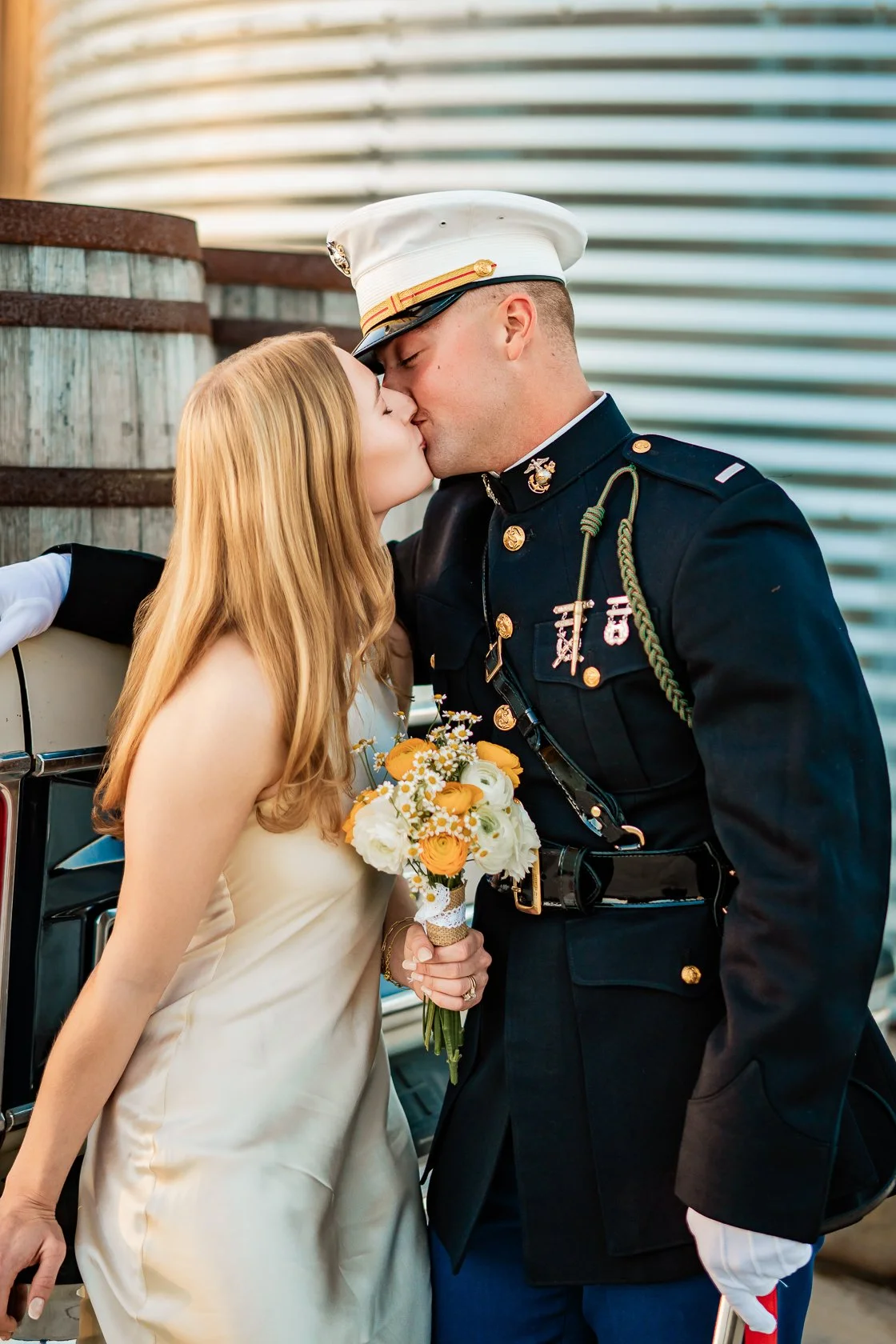 A woman in a cream-colored wedding dress kisses a man in a U.S. military dress uniform; she holds a bouquet of white, yellow, and orange flowers.
