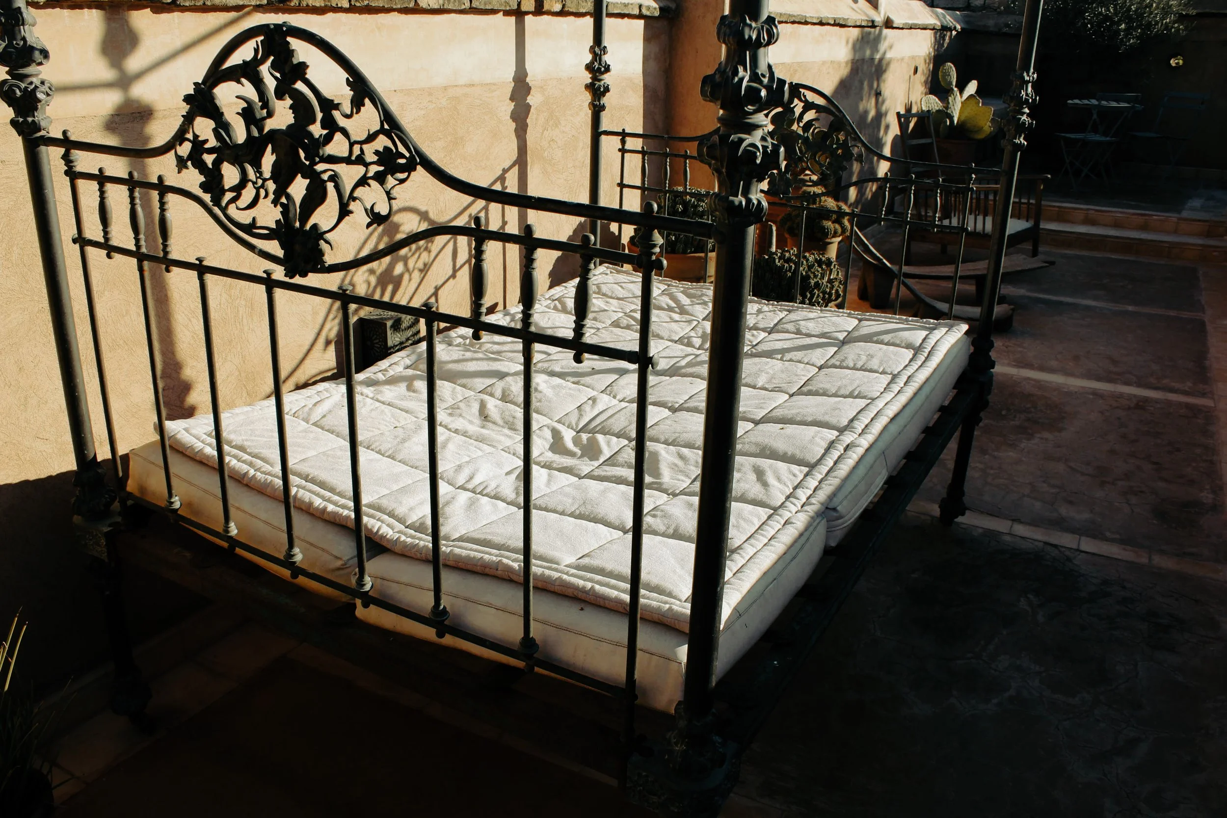 An outdoor vintage metal bed frame with a quilted mattress and no bedding, positioned on a stone patio, with a wall and potted plants in the background.