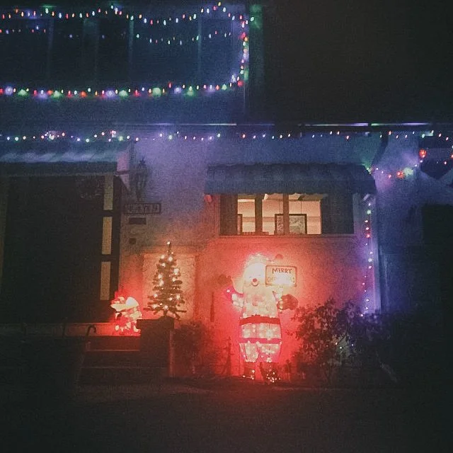 House decorated with colorful Christmas lights, inflatable Santa Claus, a small Christmas tree, and a Santa figure with a "Merry Christmas" sign above it, illuminated at night.