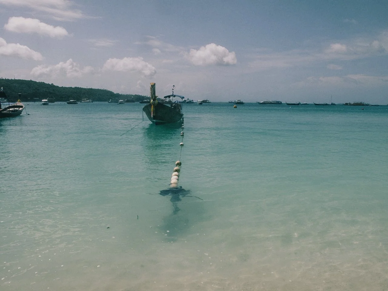 A boat anchored in clear turquoise water near a beach with several other boats in the background under a partly cloudy sky.