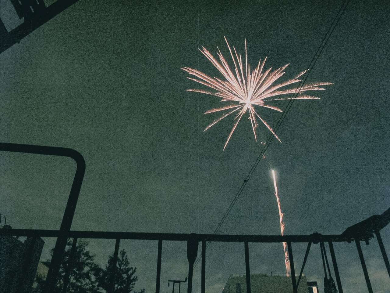 Fireworks exploding in the night sky above rooftops and trees, viewed from a balcony or patio with a metal railing.