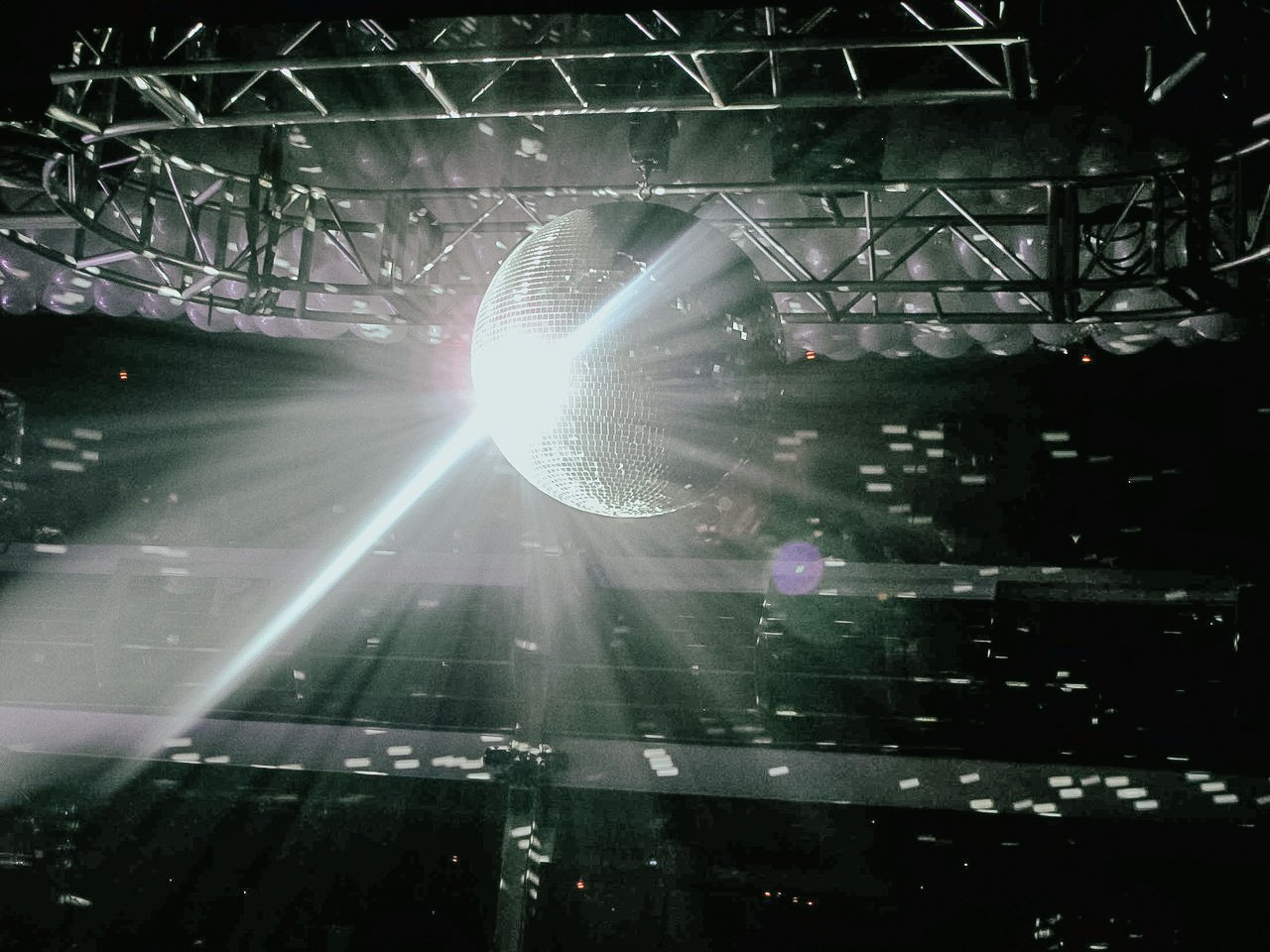 A large disco ball hanging from a metal truss with a bright light reflecting off it, creating a beam of light and scattered reflections in a dark room.