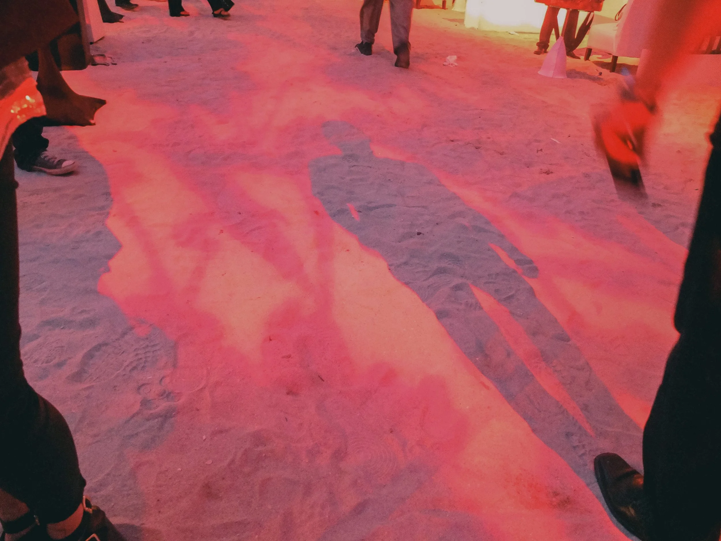 Shadow of a person walking on sandy ground, illuminated by pink and orange lighting, with other people’s legs visible around.