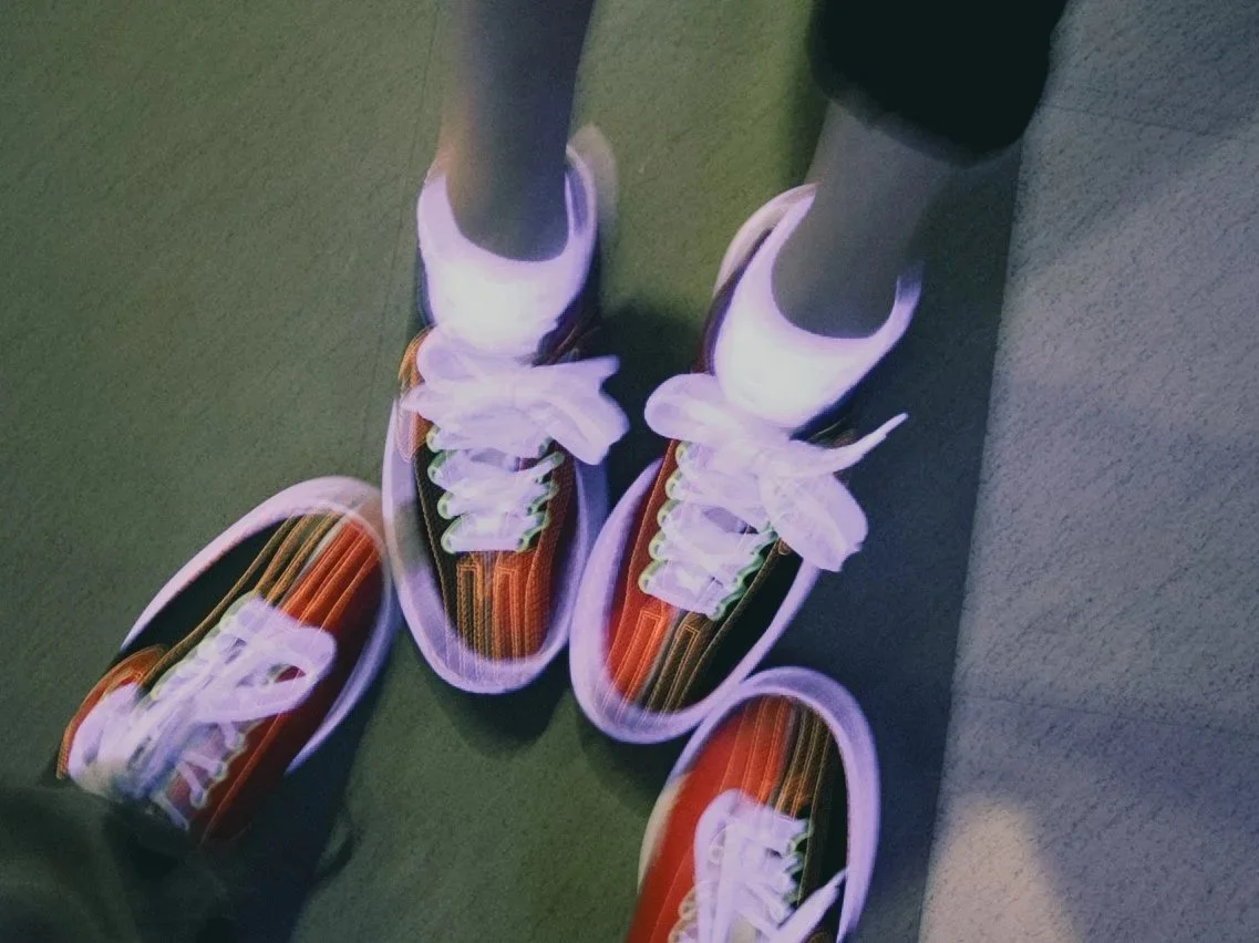 Close-up of a person's feet wearing colorful athletic shoes with white laces, standing on a gray carpeted surface next to a darker-colored area.
