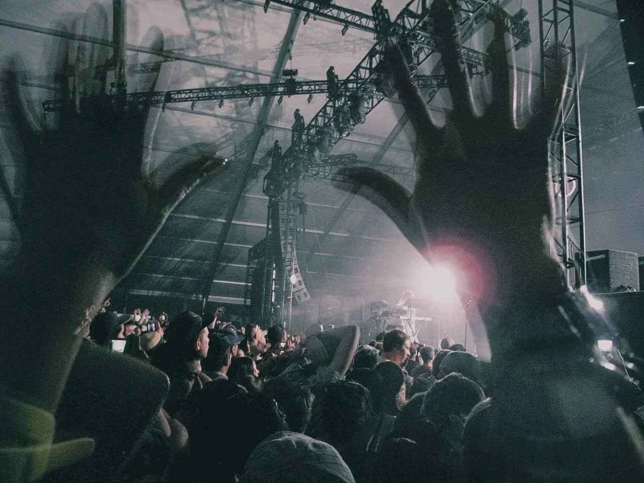 Crowd at concert with hands raised, stage with bright lights and scaffolding, DJ or performer on stage, inside a large indoor venue.