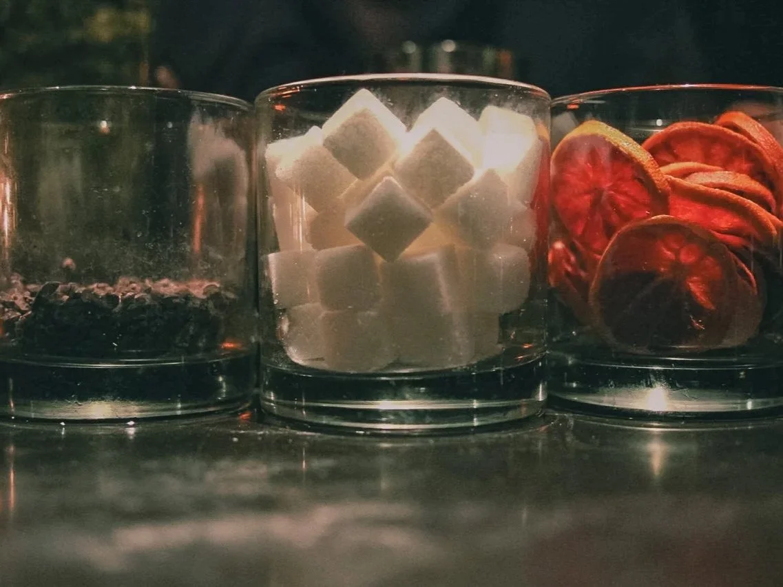 Three glass containers with different items: the left container has dark-colored small objects, the middle container has white sugar cubes, the right container has dried red tomato slices.