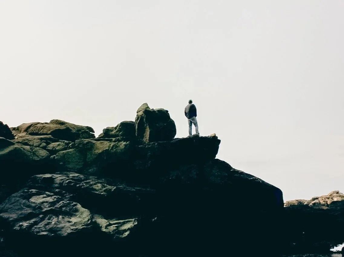 A person standing on a rocky cliff, overlooking the ocean under a cloudy sky.