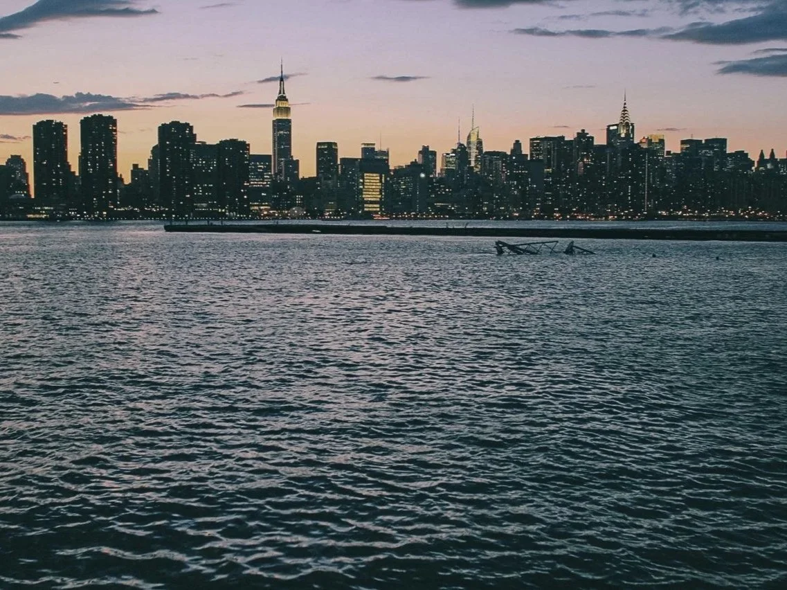New York City skyline at dusk with water in the foreground and the Empire State Building visible.