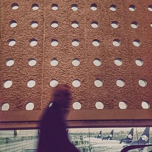 A blurred black cat sitting in front of a textured wall with circular holes, with an airport scene visible through the window below.