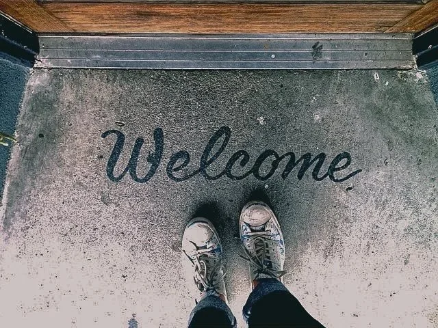 Person in sneakers and jeans standing on a concrete sidewalk with a welcome mat reading "Welcome" at the threshold of a building.