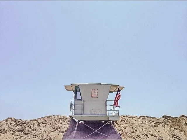 A lifeguard tower on a sandy beach with a blue sky background and an American flag hanging on the side.