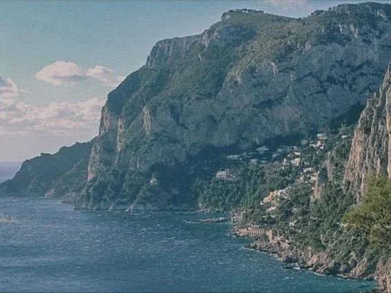 A coastal landscape with a large rocky cliff covered in greenery, overlooking a calm body of water with a few clouds in the sky.