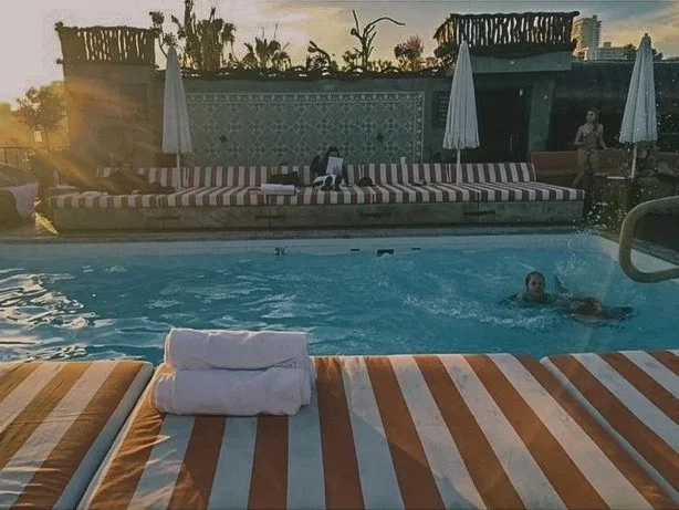 A swimming pool area at sunset with lounge chairs and umbrellas; a person swimming in the pool, with towels on the chairs, and a woman standing near the pool.