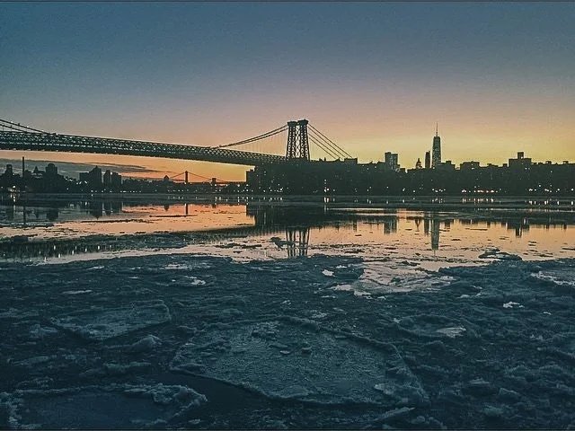 Sunset view of a city skyline with a large suspension bridge over a partially frozen river