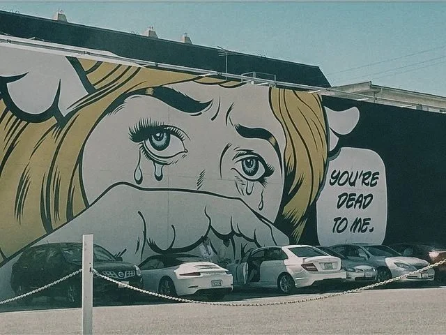A large mural of a crying woman with tears streaming down her face, covering her mouth with her hand, and a speech bubble that reads 'You're dead to me.' parked cars in front of the mural.