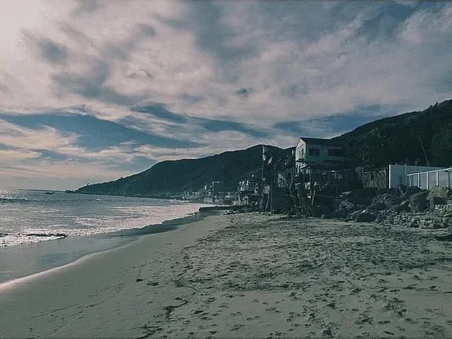 A sandy beach with ocean waves and houses built on a hill overlooking the sea, under a cloudy sky.