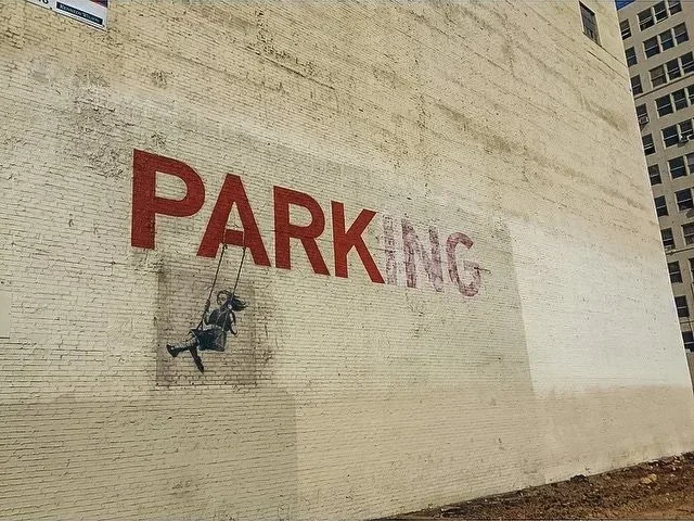 Street art of a girl on a swing hanging from the letter 'P' in the word 'PARKING' painted on a brick wall.