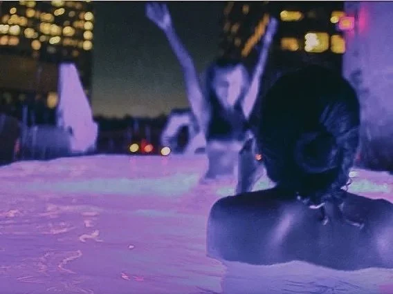 A woman sitting in a hot tub at night with city lights in the background, and another woman standing with arms raised in the background.