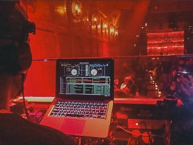 A DJ wearing headphones working at a mixing console with a laptop displaying DJ software, in a nightclub with red lighting and a crowd of people in the background.