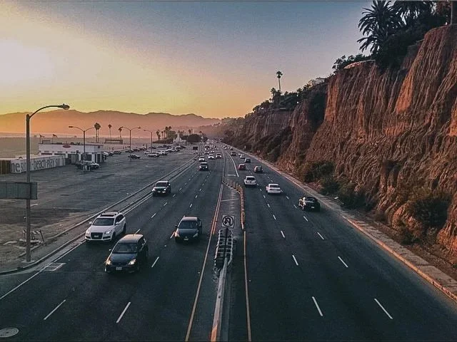 A view of a highway during sunset with cars driving in both directions, a rocky hillside on the right, and a shopping mall on the left with palm trees along the road.