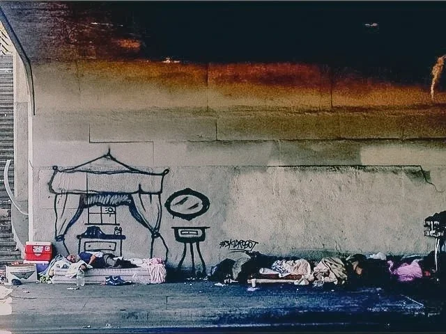 Homeless people sleeping under a bridge with graffiti on the wall behind them.
