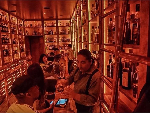 People inside a warm-lit bar or liquor store with shelves full of bottles of alcohol on the walls, engaged in conversations and socializing.