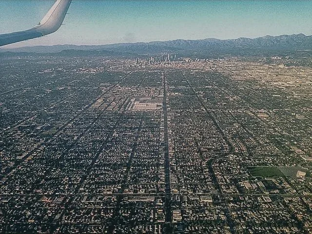 View of a city from an airplane window, showing urban landscape, grid-like streets, and mountains in the distance.