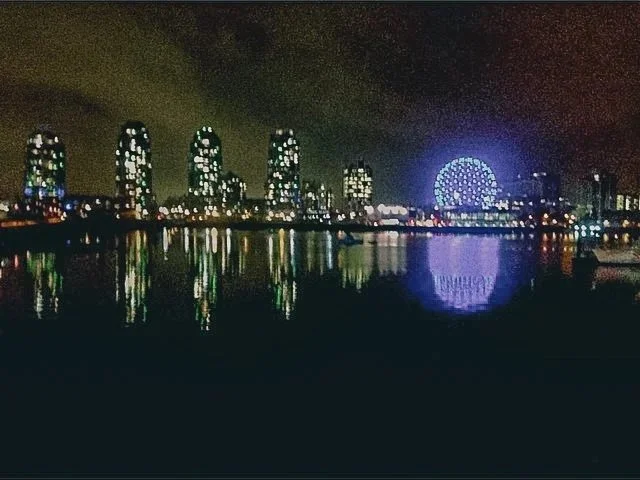 Nighttime city skyline with illuminated skyscrapers and a bright Ferris wheel reflected on the water.