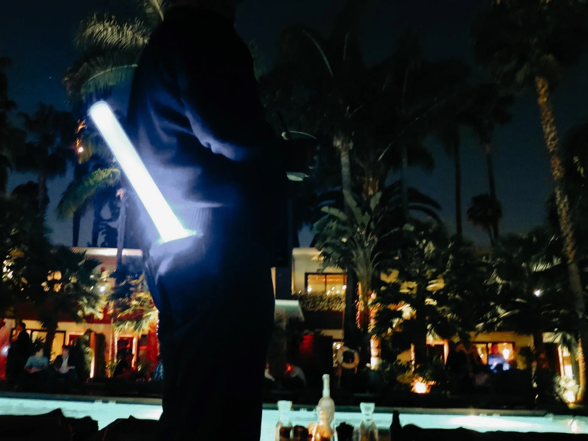 A person at night holding a glowing light stick in front of palm trees and a pool with drinks and people in the background.