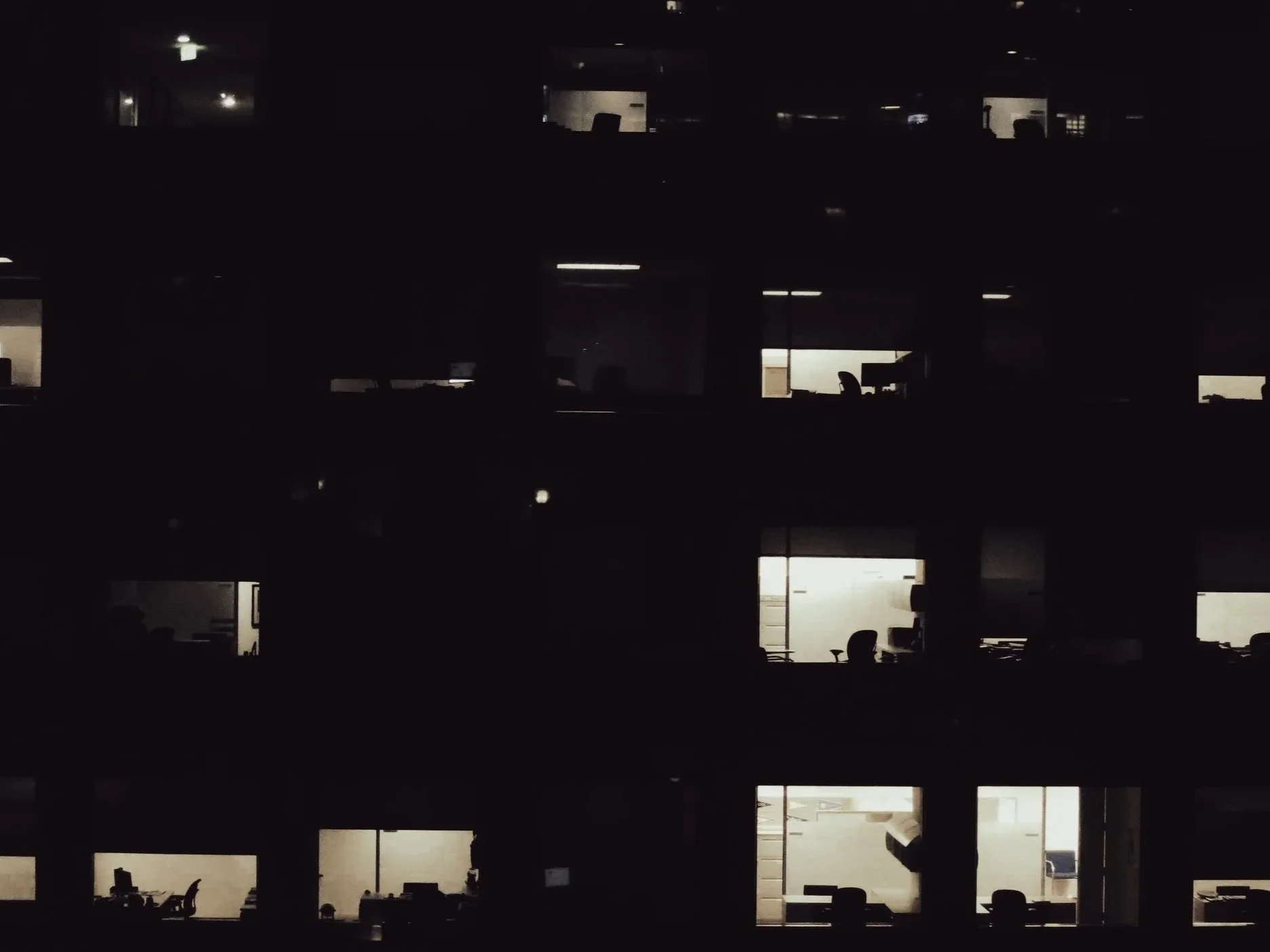 Dark multi-story office building at night, with lit office windows showing desks and chairs inside.