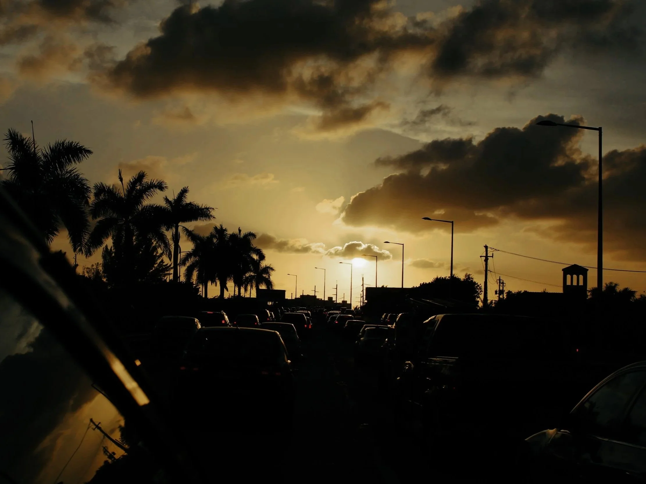 Sunset over a parking lot with silhouettes of palm trees, streetlights, and parked cars against a cloudy sky.