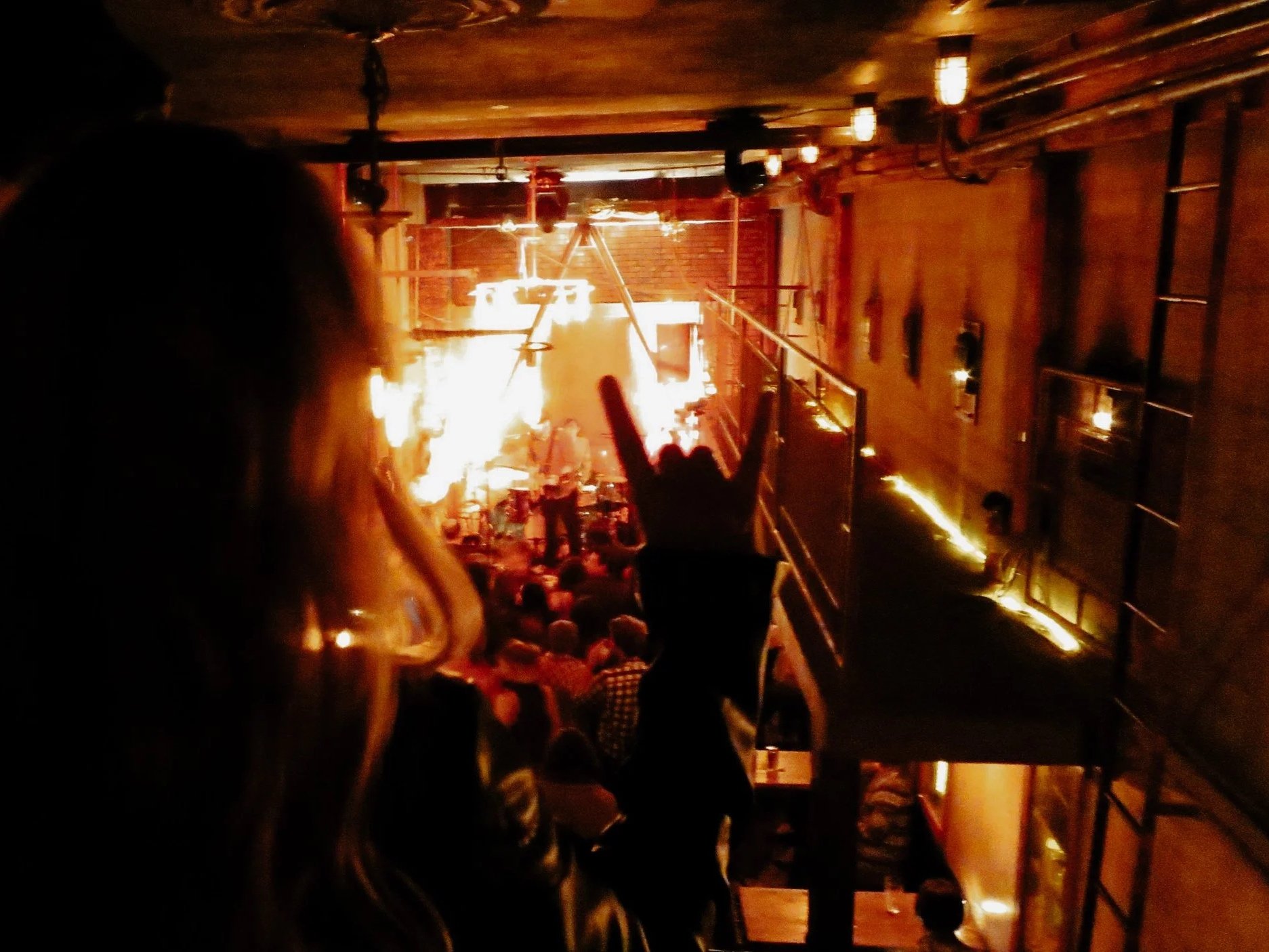 A concert or live music performance with a stage illuminated by bright lights, and an audience watching from a balcony in an industrial-style venue with exposed brick walls and metal railings.