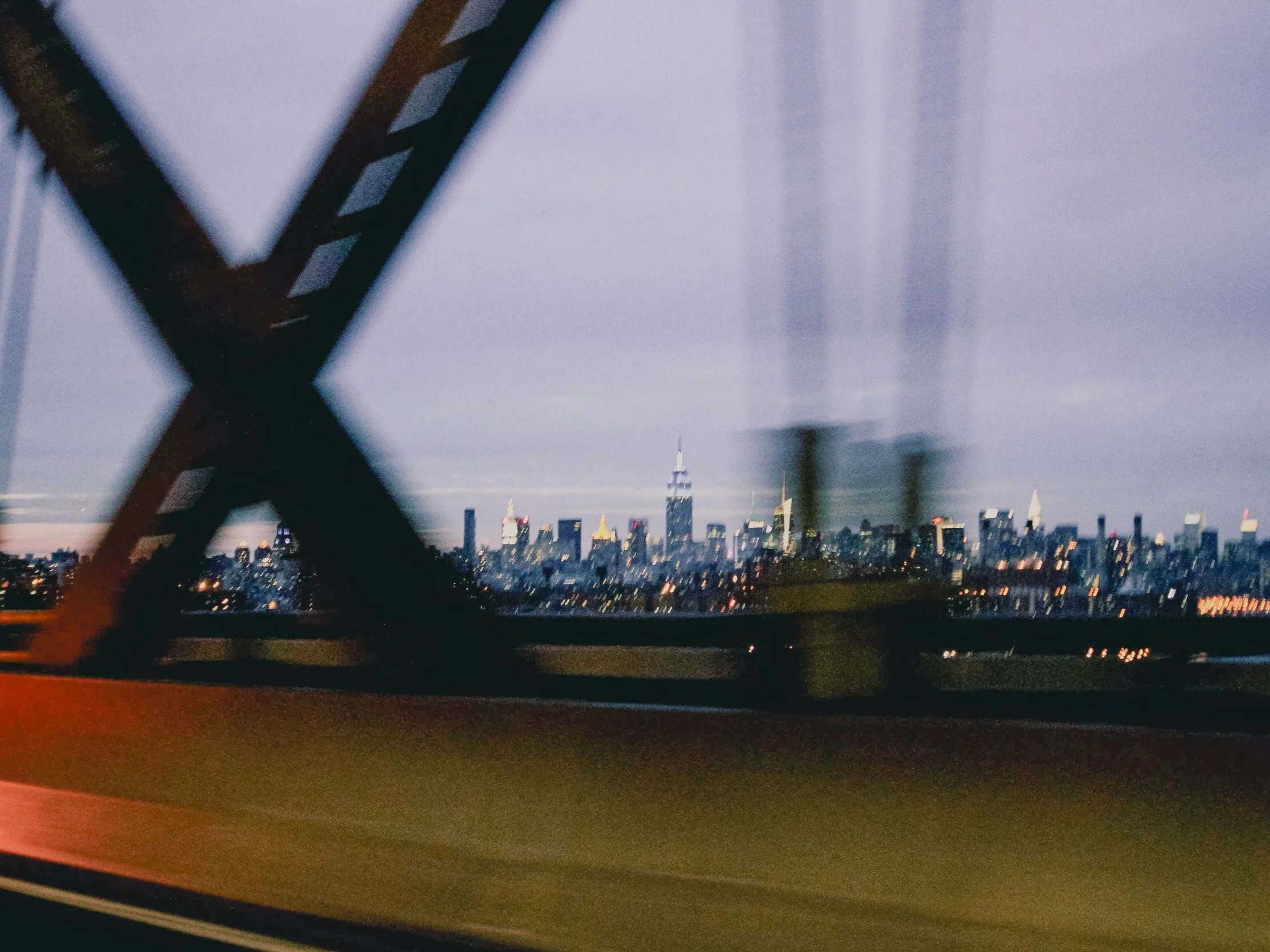 City skyline at dusk seen through a the structure of a bridge or building, with a blurred, cloudy sky and illuminated skyscrapers in the background.