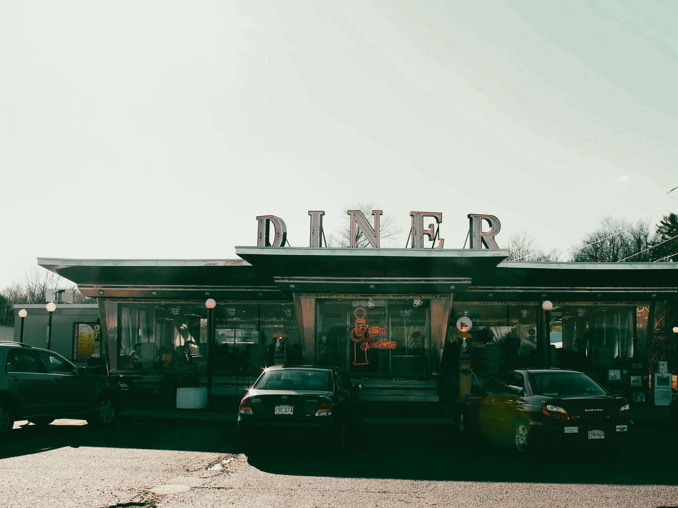 Front view of a retro-style diner with large illuminated 'DINER' sign on the roof, cars parked in front, and a neon sign inside reading 'Miami Garden'.