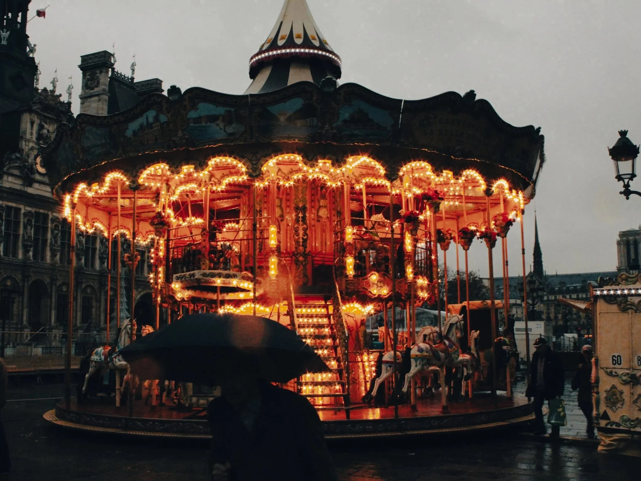 A brightly lit carousel with ornate decorations and carousel horses, set against a cloudy sky and historic buildings in the background. A person holding an umbrella is in the foreground.