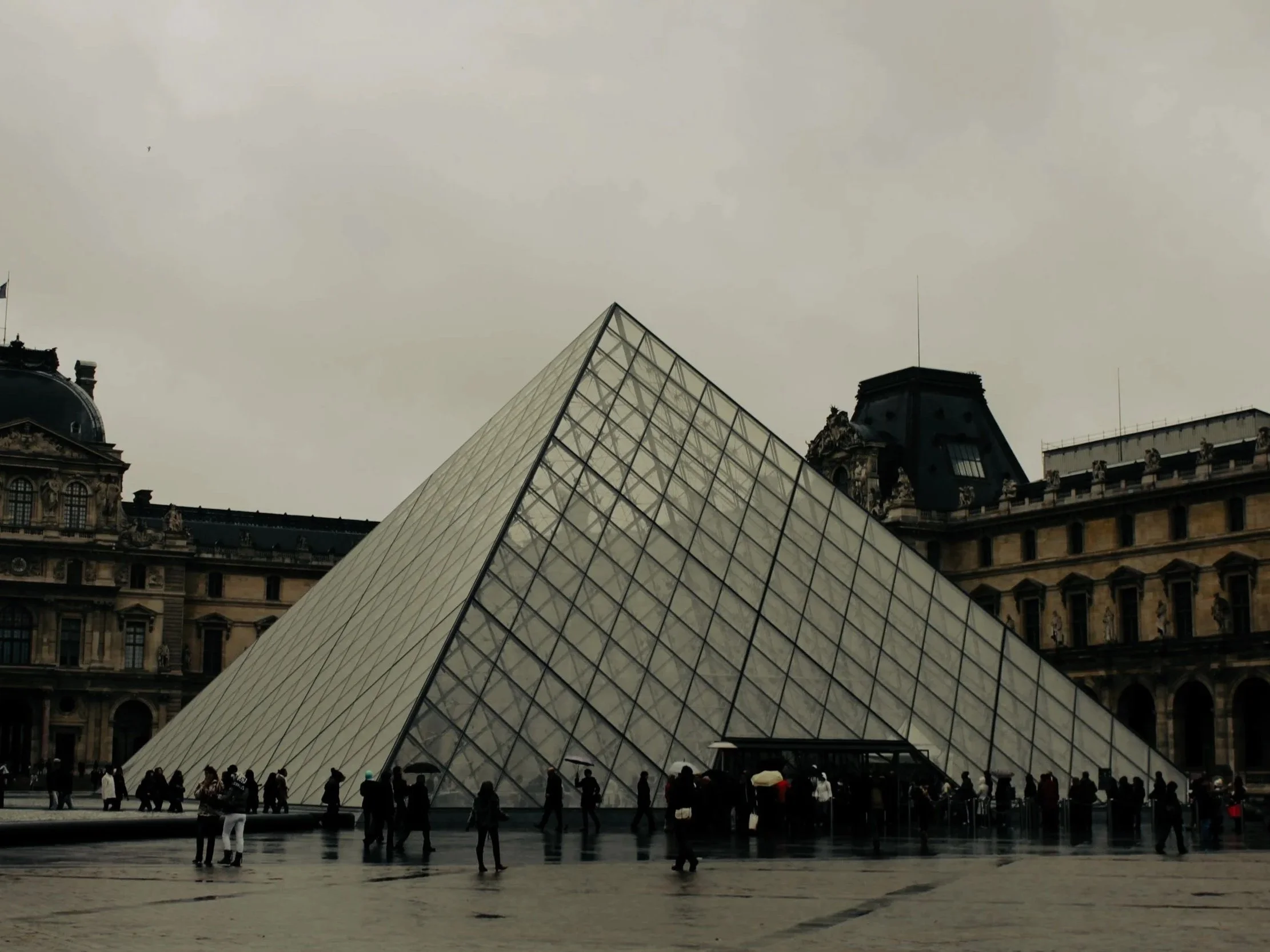The Louvre Pyramid in Paris, France, with visitors walking around on a rainy day under umbrellas.