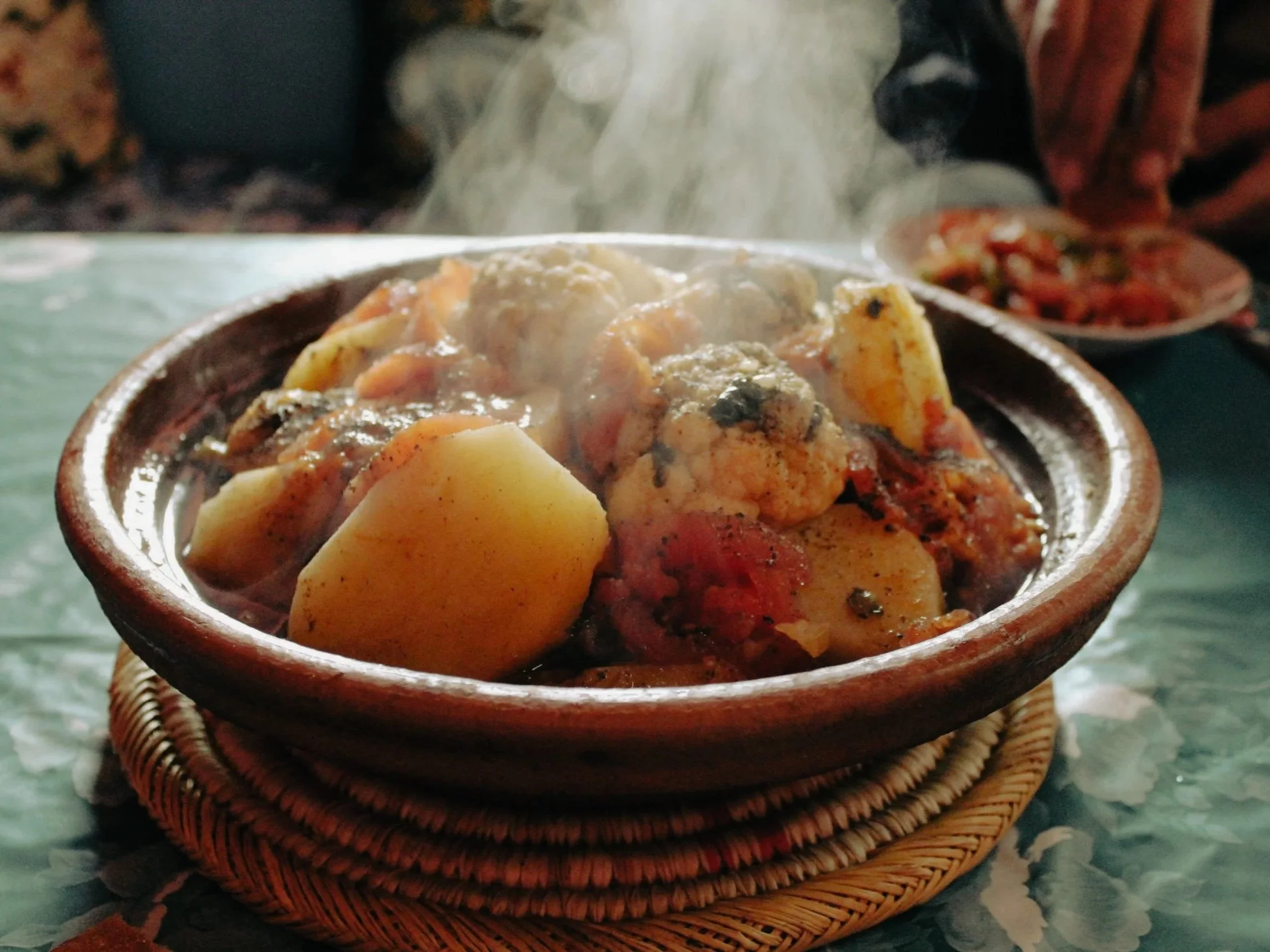 Steaming bowl of traditional food with potatoes and vegetables garnished with spices on a woven trivet, with a person in the background.