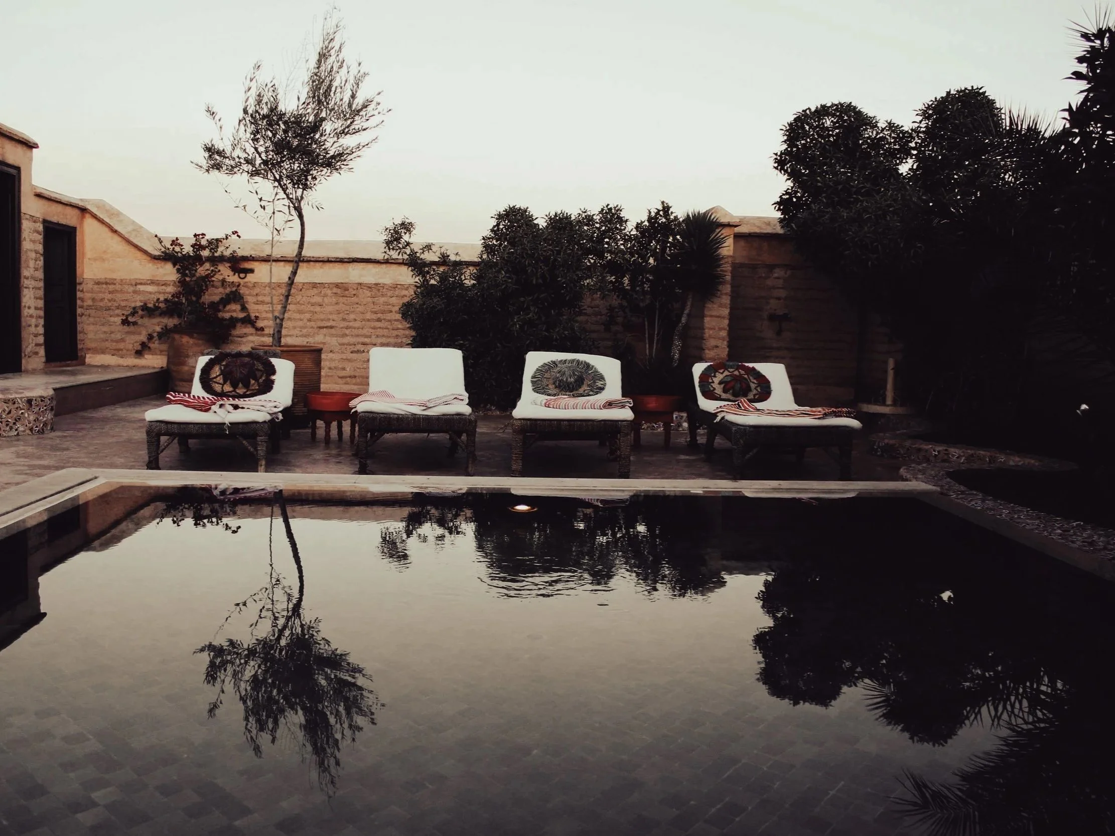 A backyard scene during dusk with four white lounge chairs with colorful pillows and striped towels near a rectangular swimming pool. There are potted plants and trees, with a brick wall in the background and the pool's reflection visible in the wate