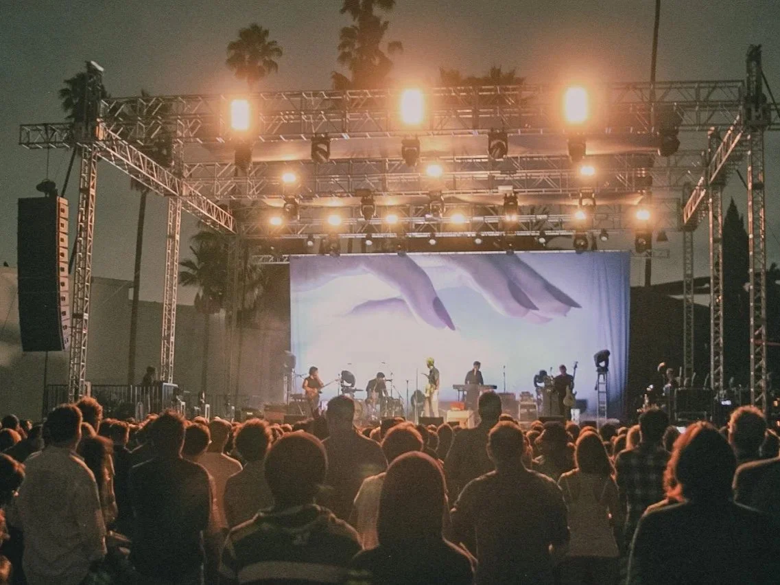 Outdoor concert stage with musicians performing, crowd watching, trees in background, stage lights shining.