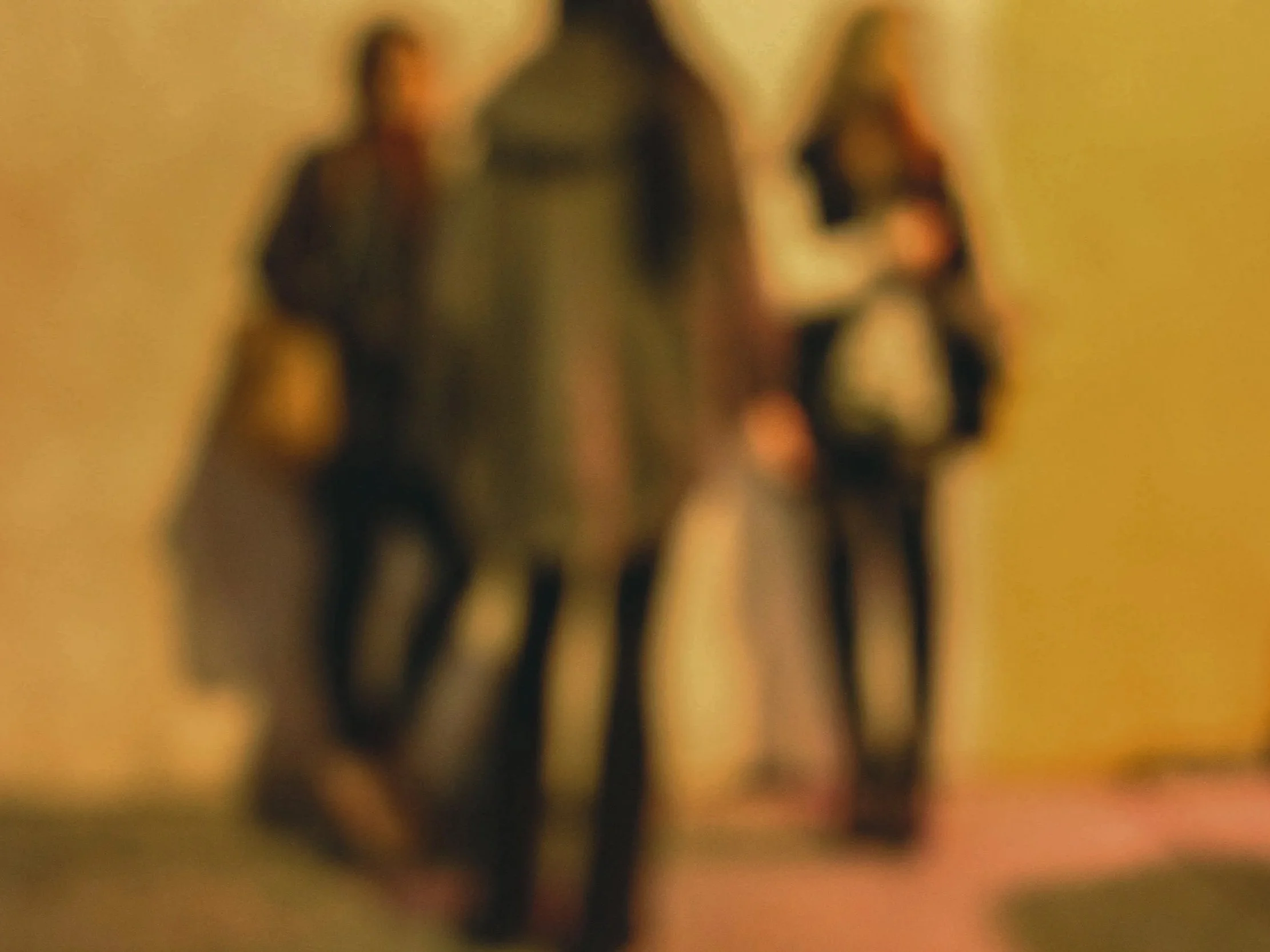 Three blurry individuals standing in a hallway or room, one in the foreground and two in the background.