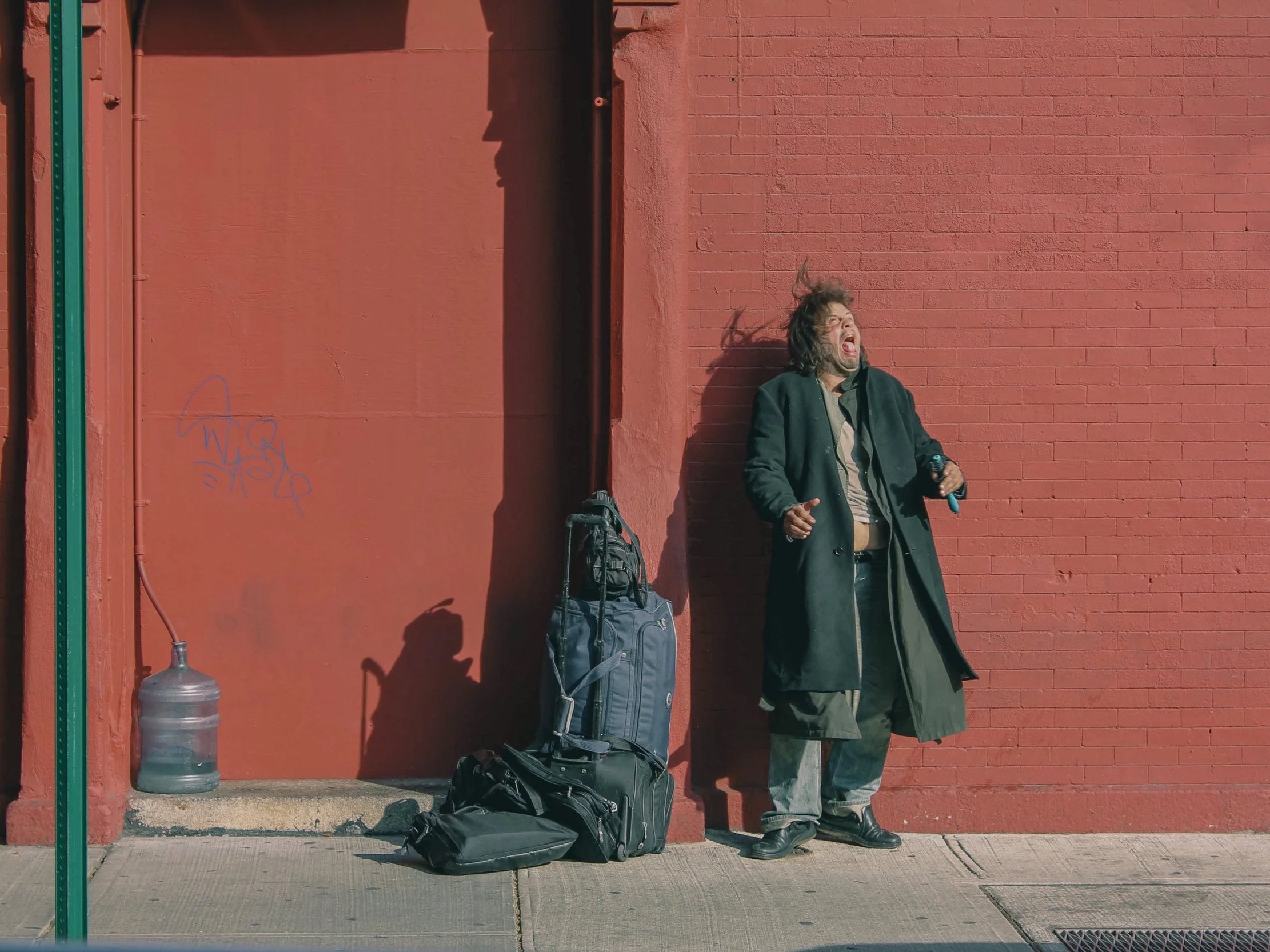 A man with long hair and beard singing or shouting on the sidewalk against a red brick wall, with luggage and a water jug nearby.