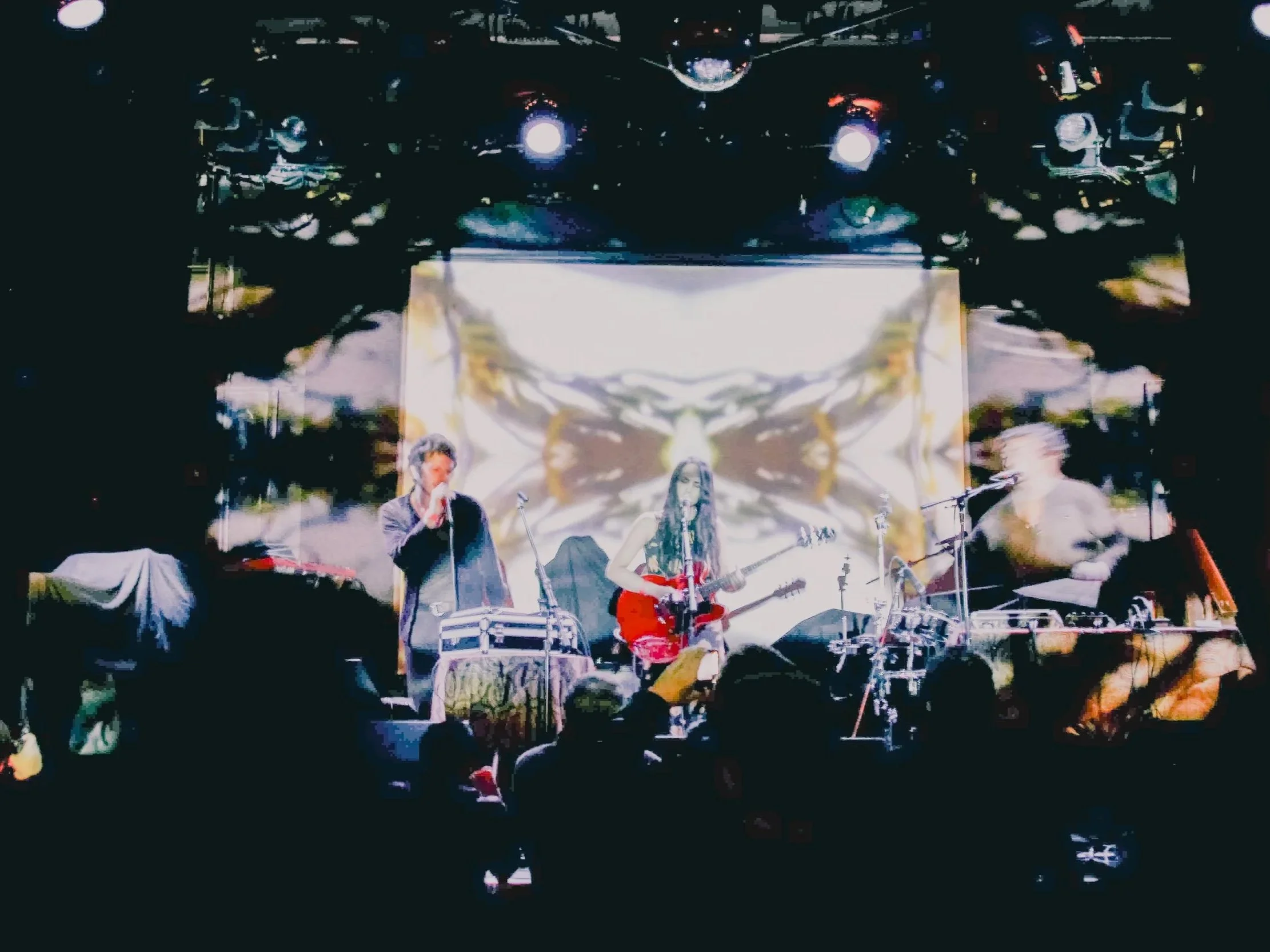 A live music performance on stage with three musicians: a singer on the left, a guitarist in the center, and a drummer on the right. The stage features colorful lighting and a digital backdrop projection.