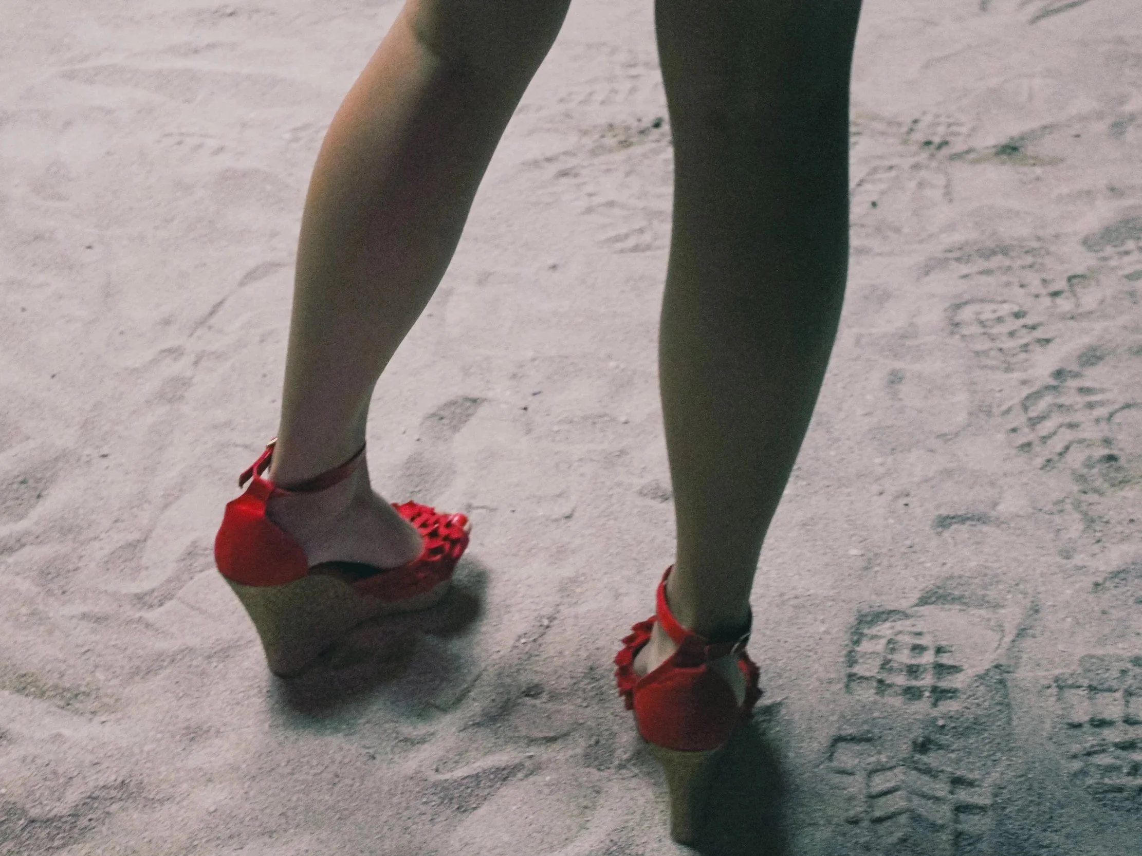 A person's legs and red high-heeled shoes on sandy ground with footprints.
