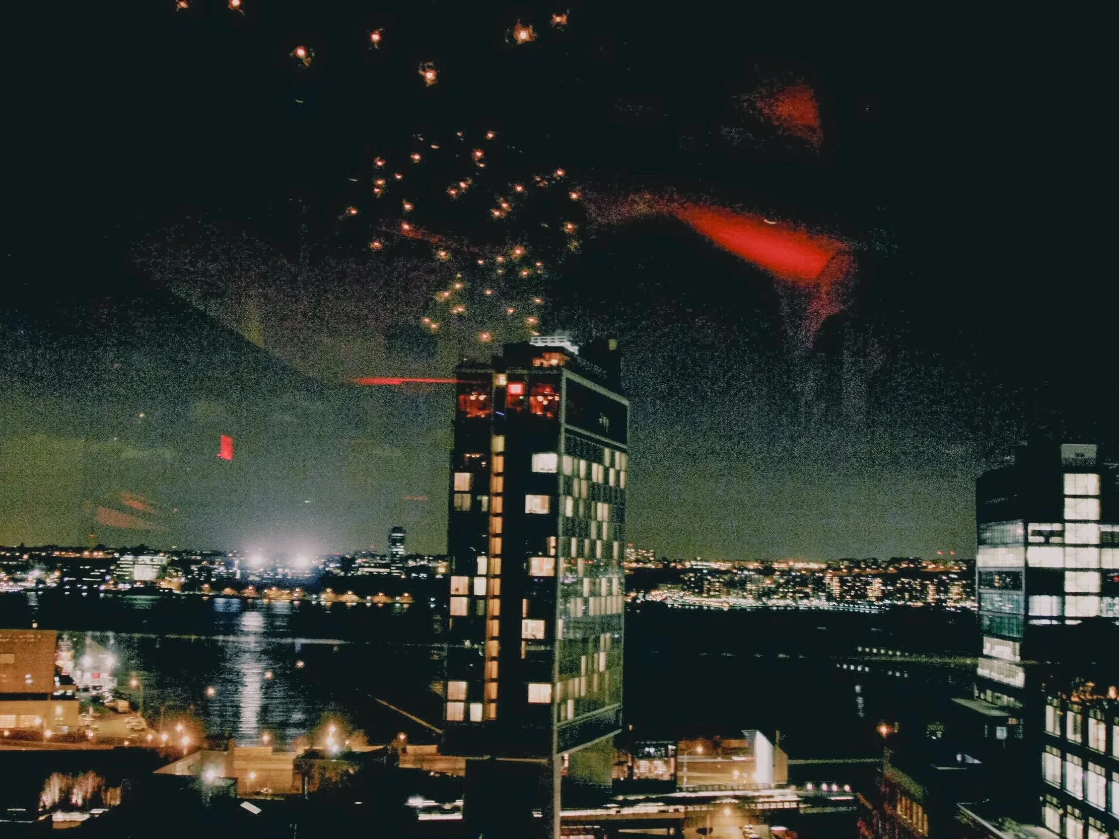 Nighttime cityscape with illuminated buildings, fireworks in the sky, and a river reflecting city lights.