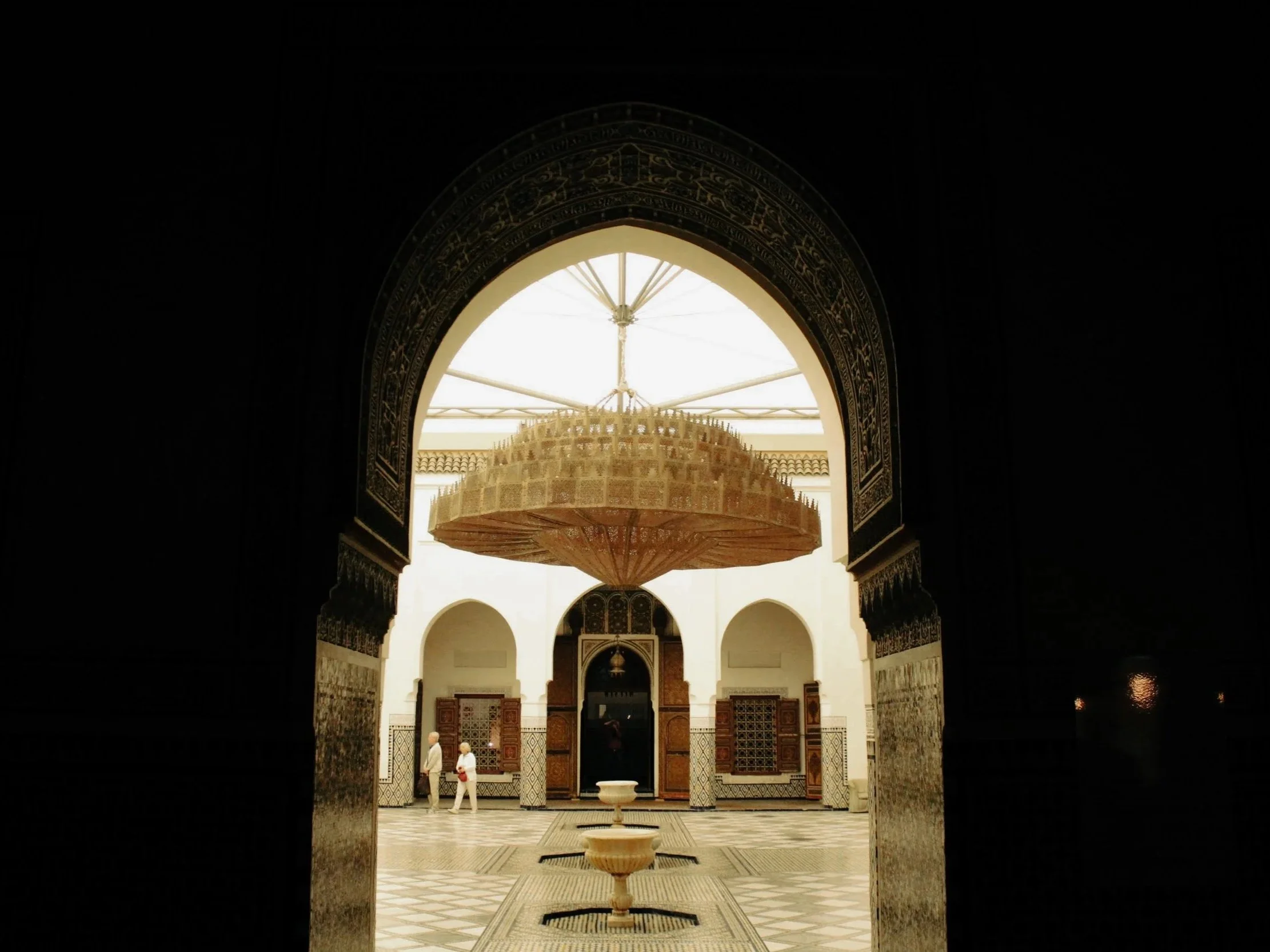 Interior view of a traditional Moroccan riad with a central fountain, wooden chandelier, arched doorways, and decorative tiles, seen through a dark doorway.