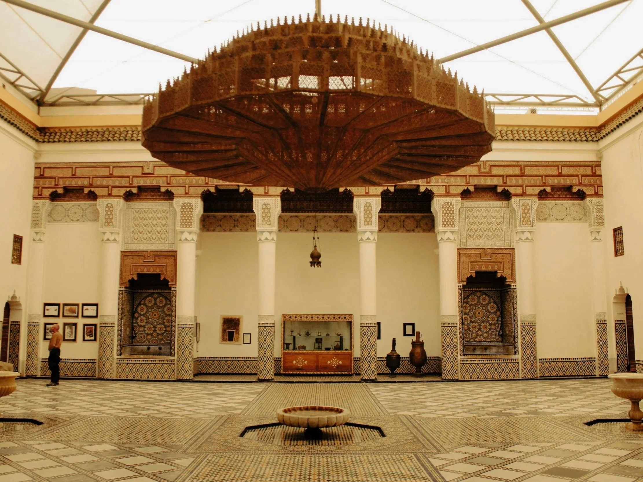 An ornate indoor courtyard with patterned tile floors, white columns, and decorated walls. A large, circular wooden chandelier hangs from the ceiling, and there are small displays and decorative vases along the walls.