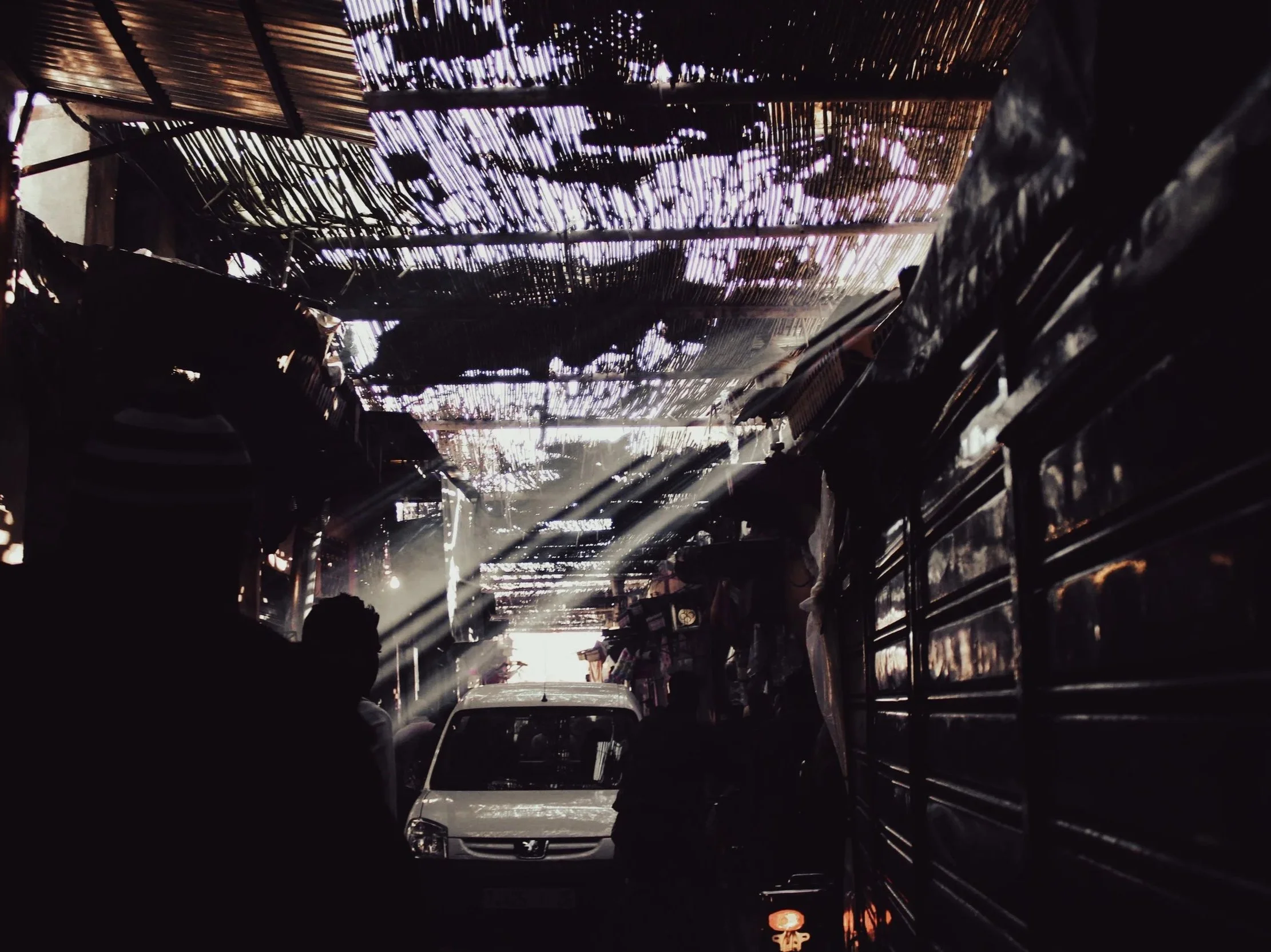 A street scene with a white car, several people, and makeshift stalls. Sunlight filters through a roof made of woven bamboo or similar material, casting shadows.