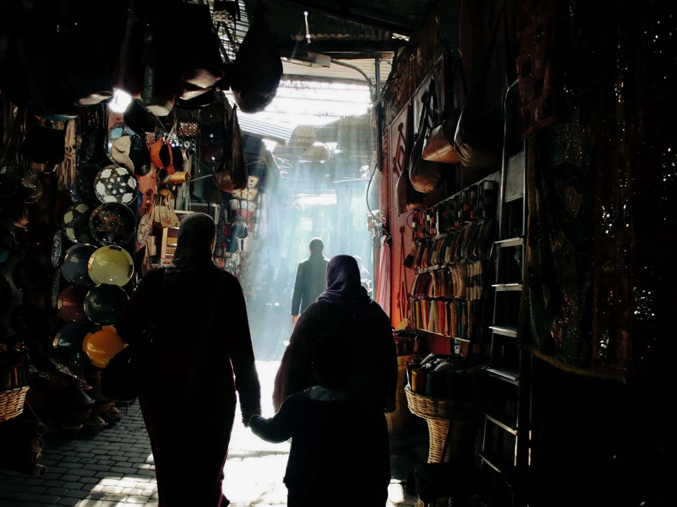 A family holding hands inside a narrow market, walking towards bright sunlight outside, surrounded by shelves of hats, plates, and textiles in a market shop.