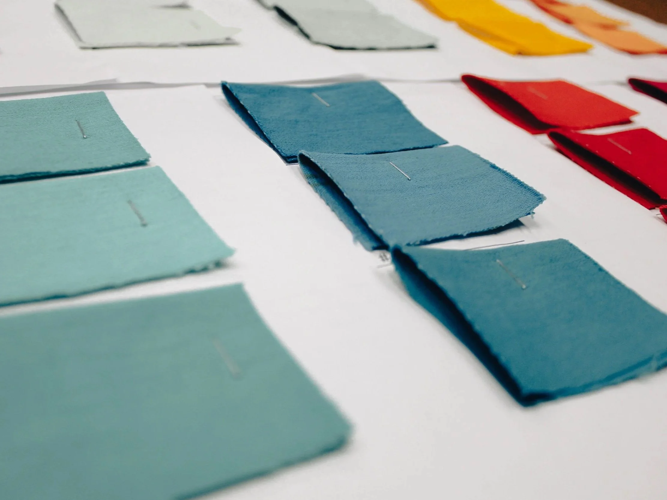 Color swatches of fabric in shades of green, blue, red, yellow, and white displayed on a white surface, with some swatches pinned with straight pins.