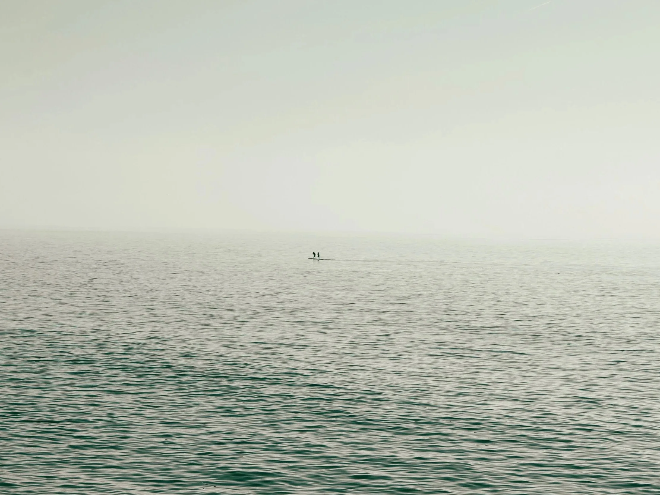 Two small figures on a boat in a vast, calm, and misty body of water with a hazy horizon.